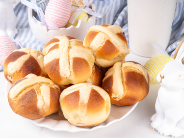 Plate of traditional hot cross buns surrounded by decorated Easter eggs and spring-themed decorations.